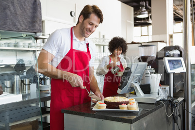 Smiling waiter slicing cake with waitress behind him