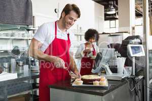 Smiling waiter slicing cake with waitress behind him