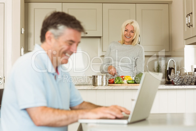 Happy mature man using laptop while wife prepares vegetables