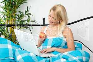 Frau sitzt mit Laptop im Bett und isst einen Apfel