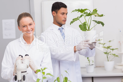 Scientist smiling at camera while colleague looking at plant