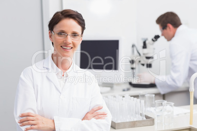 Scientist smiling at camera arms crossed and another working wit