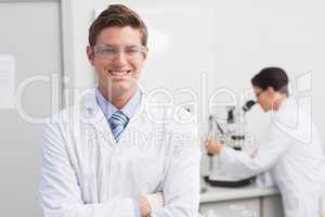 Scientist smiling at camera arms crossed and another working wit