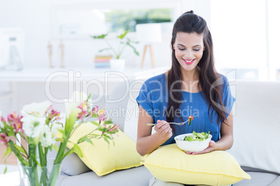 Smiling beautiful brunette sitting on the couch and eating salad