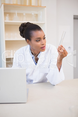Scientist looking at test tube while using laptop