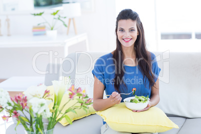 Smiling beautiful brunette sitting on the couch and eating salad