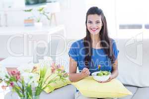 Smiling beautiful brunette sitting on the couch and eating salad