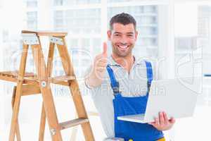 Smiling handyman with laptop gesturing thumbs up in office