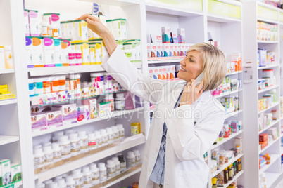 Smiling pharmacist phoning and taking medicine from shelf