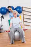 Therapist helping senior woman fit dumbbells on exercise ball