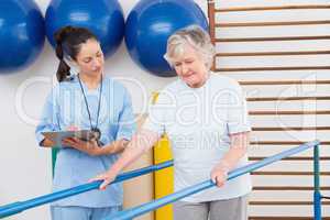 Therapist looking senior woman walking with parallel bars
