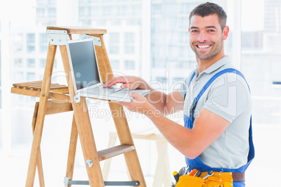 Smiling handyman using laptop by ladder in office