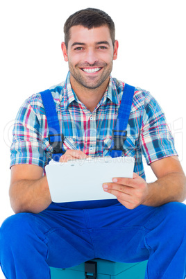 Happy plumber writing on clipboard while sitting on toolbox