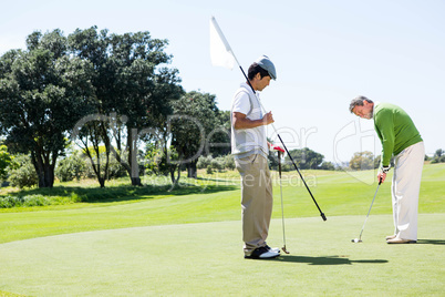 Golfer holding hole flag for friend putting ball