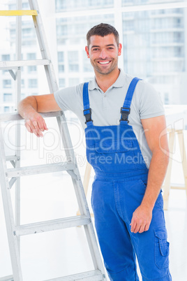 Happy handyman in overalls leaning on ladder at office
