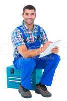 Happy plumber writing on clipboard while sitting on toolbox