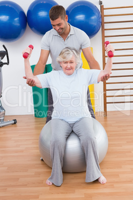 Trainer helping senior woman lift dumbbells on exercise ball