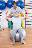 Trainer helping senior woman lift dumbbells on exercise ball