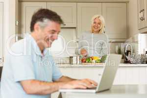Happy mature man using laptop while wife prepares vegetables