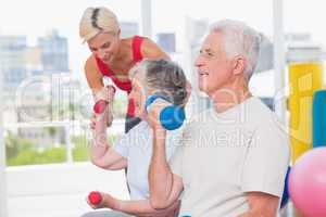 Senior man lifting dumbbells while trainer assisting woman at gy