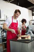 Smiling waiter slicing cake with waitress behind him