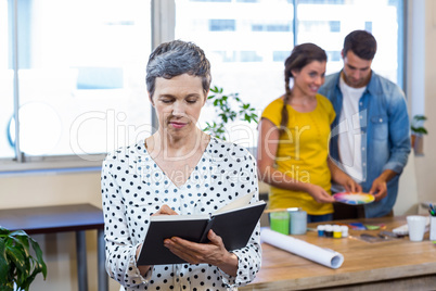 Casual businesswoman writing in diary while colleagues working