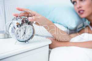 Sleepy young woman in bed extending hand to alarm clock