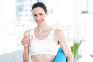 Smiling brunette holding exercise mat and glass of water