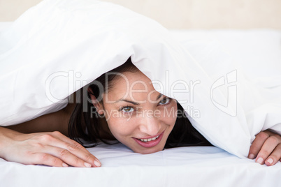 Pretty brunette looking at camera and hiding under the duvet