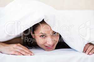 Pretty brunette looking at camera and hiding under the duvet