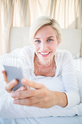 Smiling blonde woman lying on the bed and texting with her mobil
