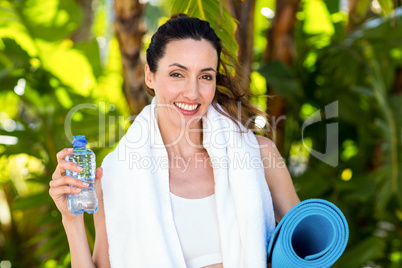 Smiling brunette holding bottle of water and exercise mat