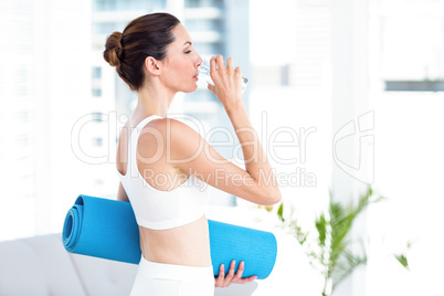 Brunette drinking water while holding exercise mat