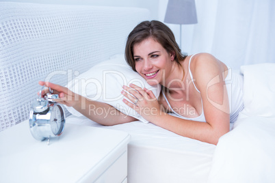 Happy brunette extending hand to alarm to clock in bed