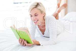 Smiling blonde woman lying on the bed and reading a book