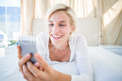 Smiling blonde woman lying on the bed and texting with her mobil