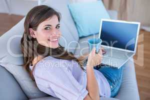 Pretty brunette using her laptop with cup of coffee on couch