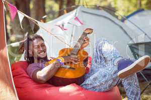 Happy hipster relaxing on campsite playing guitar