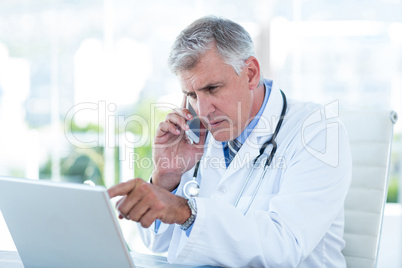 Serious doctor working on laptop and having phone call