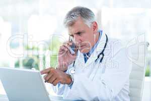 Serious doctor working on laptop and having phone call