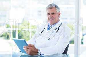 Smiling doctor sitting at his desk and holding tablet