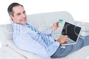 Smiling businessman lying on a sofa holding a card and laptop