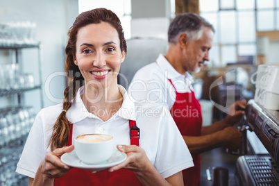 Smiling barista holding a cup of coffee with colleague behind
