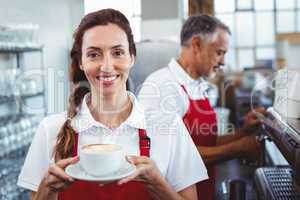 Smiling barista holding a cup of coffee with colleague behind