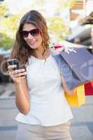 Smiling woman with sunglasses and shopping bags using her smartp