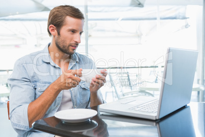 Young happy man holding his coffee while looking at his laptop Young happy man holding his coffee while looking at his laptop