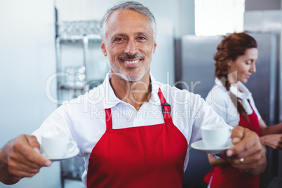 Smiling barista holding cups of coffee with colleague behind