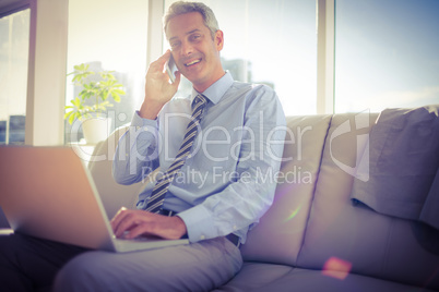 Businessman sitting on a couch having phone call and using lapto