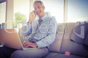 Businessman sitting on a couch having phone call and using lapto