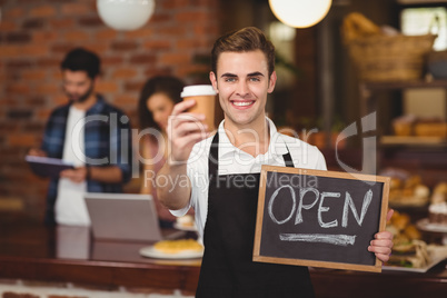 Smiling barista holding take-away cup and open sign